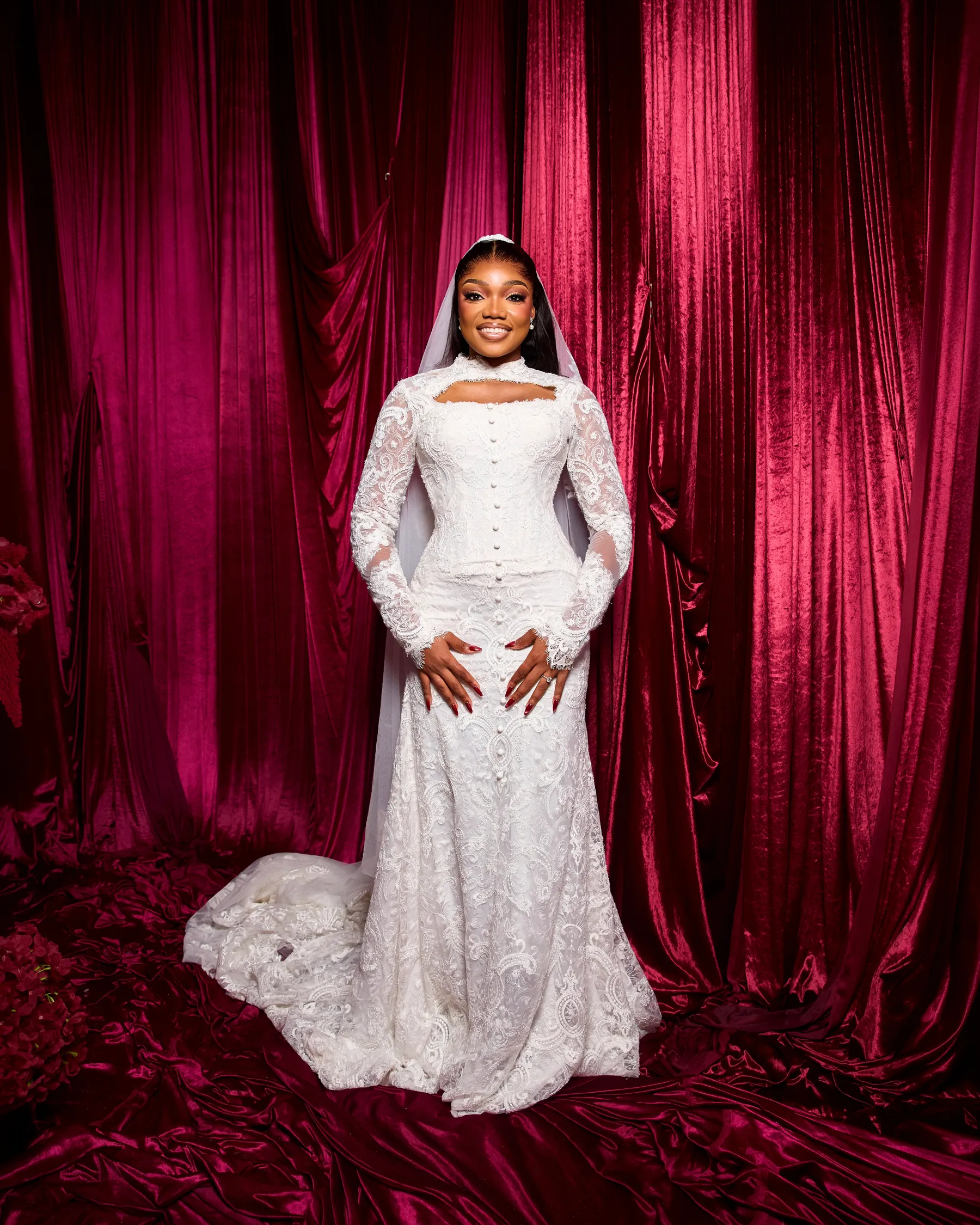 Jiofit bride in a long-sleeved lace gown with a high keyhole collar and covered buttons, its train spread over crimson velvet
