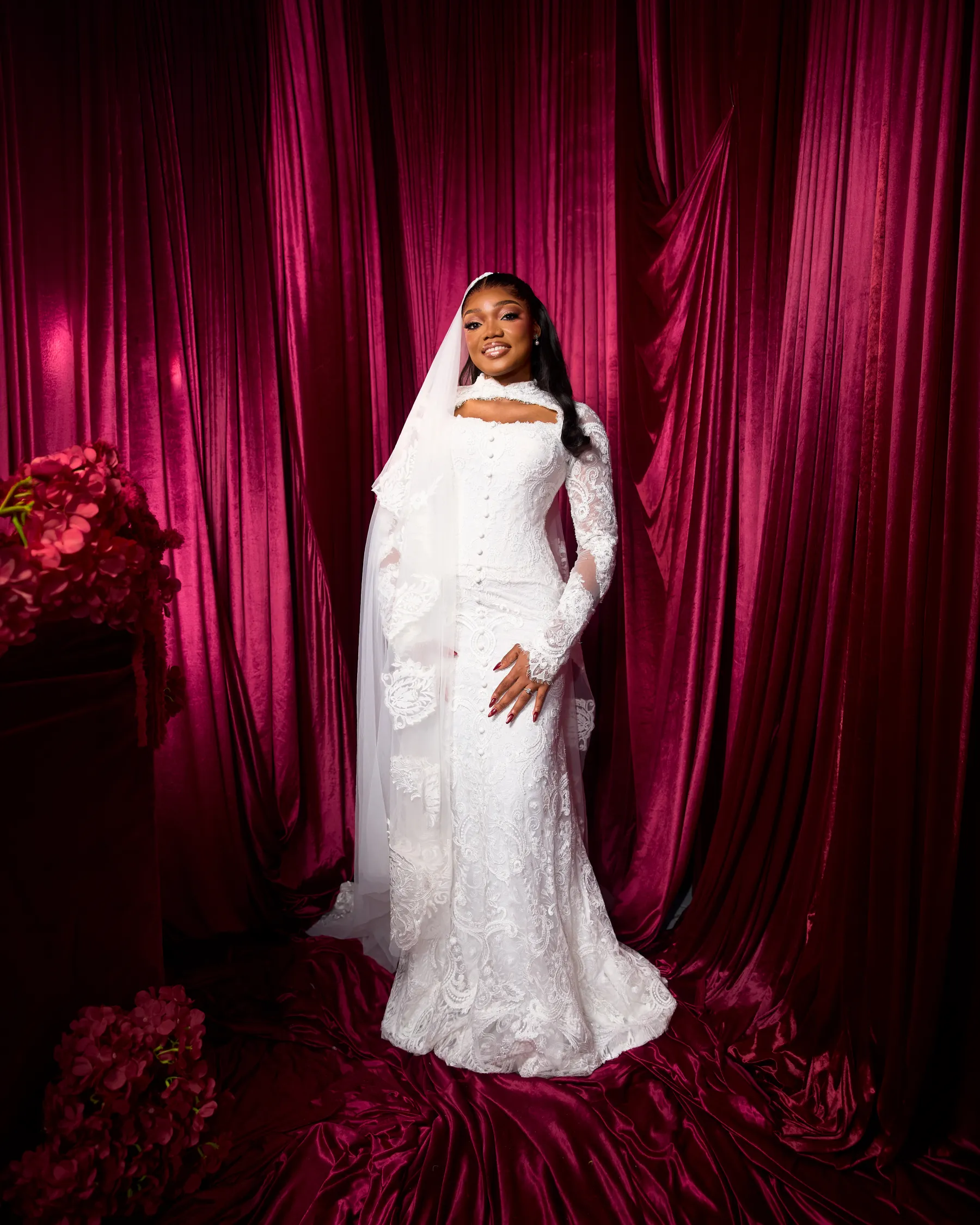 Jiofit bride in the long-sleeved lace gown, her veil drawn over one shoulder, beside red roses and crimson velvet